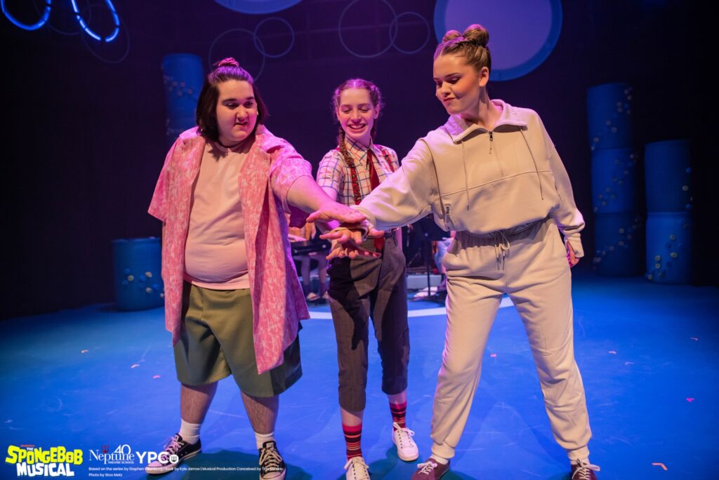 Three teenagers as the leads in SpongeBob : The Musical. They stand in a line with their palms in a hand stack. 