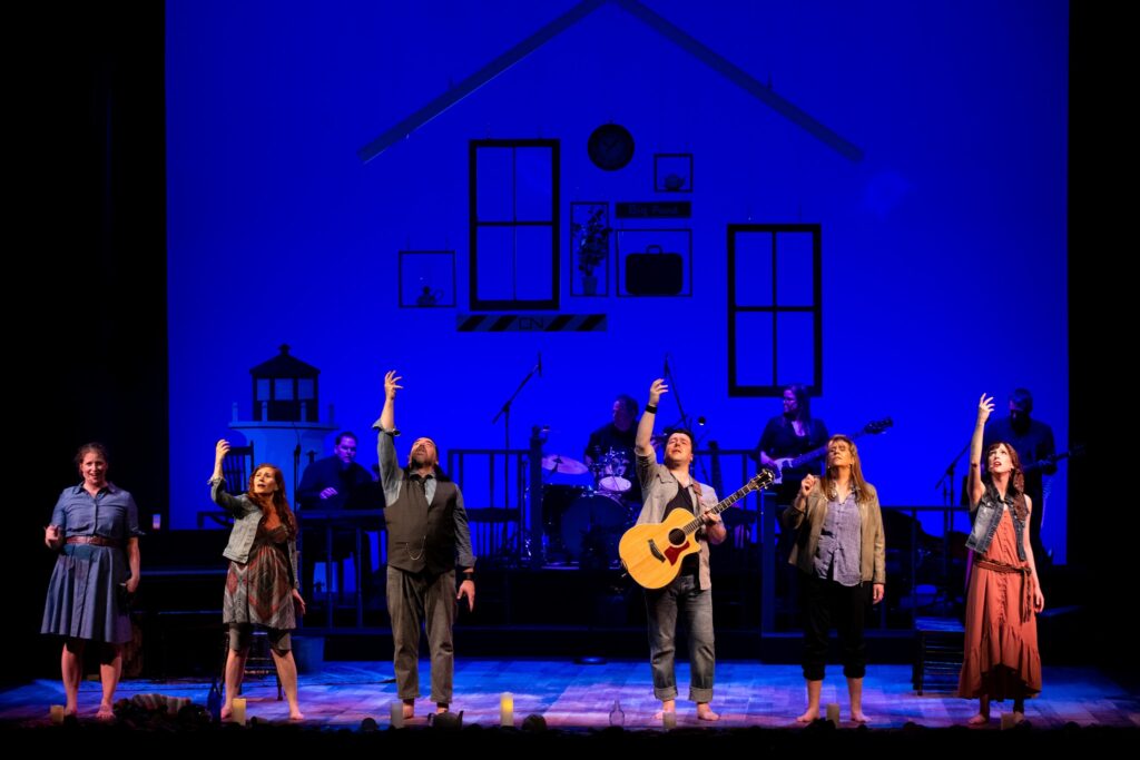 Against a blue background with a cartoon house drawn on it stand six performers in casual, modern clothes. There are four women and two men, they stand in a horizontal line in front of the stage. They all look upward, and some hold up their left arms. One of the men has a guitar in his hand. You can see the outlines of four musicians in shadow behind them. 