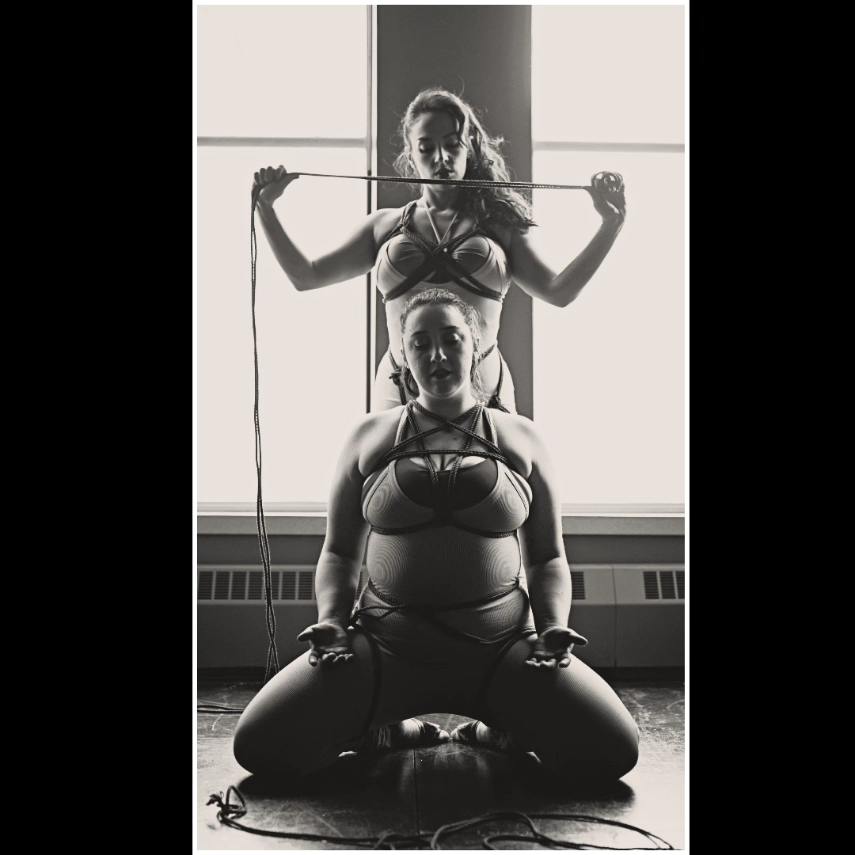 A black and white photo of two women in bikini tops and pants. One sits in front of the other. The one in the back is standing over her partner suggestively holding a long piece of rope. Both women have ropes crisscrossing over their chests as well. These women are lovers.