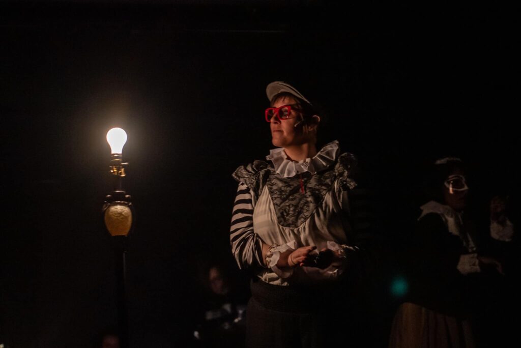 A non-binary actor in a cap with red glasses and a Elizabethan Clown-adjacent shirt sits to the right of a ghost lamp on a darkened stage. 