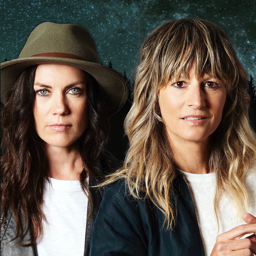 Lisa MacIsaac and Brenley MacEachern stare directly at the camera. Lisa is a white woman with brown hair, Brenley is a white woman with blonde hair. They both wear white t-shirts with black blazers over them. Lisa wears a cowboy style hat. Both have intense eyes and slight smiles.