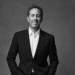 A black and white headshot of Jerry Seinfeld in a suit, looking directly at the camera and smiling.