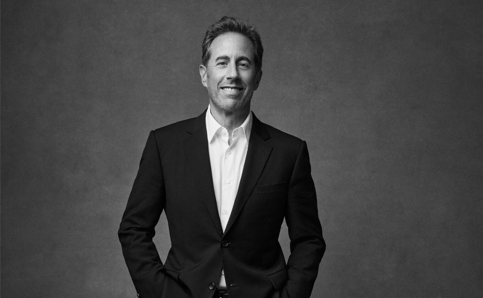 A black and white headshot of Jerry Seinfeld in a suit, looking directly at the camera and smiling.