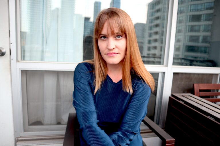 A white woman sits with her arms folded in front of her looking at the camera. She has reddish blonde hair with bangs, and an intense expression. She wears a blue sweater which sets off the colour of her hair beautifully.