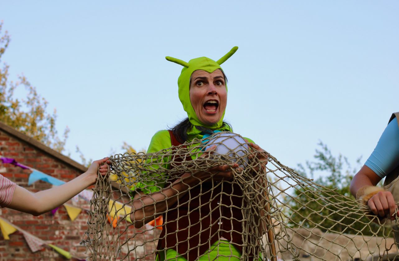 [ID: The background is a blue sky featuring the blurred Cambridge Battery. The focal point of the image is Jimmy the Cricket played by Geneviève Steele. Jimmy is caught in a net and holds a magic crystal ball.]