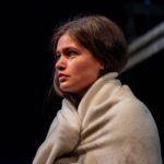 A young girl with brown hair looks very sad away from the camera, she holds an airline blanket around her.