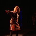 A person with red hair and wearing a crown and kilt, celebrates onstage in a golden light.