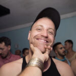 A white man in a black tank top with a black ball cap on grins at his friend who is taking his photo. His friend holds him under his chin with his hand.