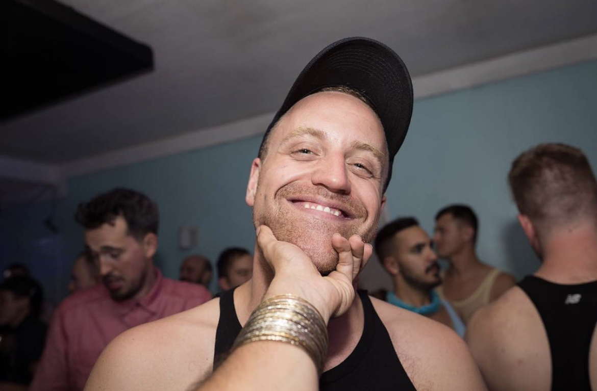 A white man in a black tank top with a black ball cap on grins at his friend who is taking his photo. His friend holds him under his chin with his hand.