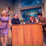 A woman and three men wearing vaguely 1960s attire inside a floral shop looking at a strange little fly-trap plant. A Black man peeks in at the plant from the back window.