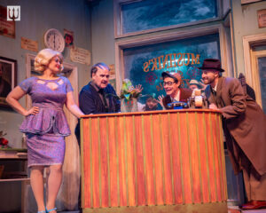 A woman and three men wearing vaguely 1960s attire inside a floral shop looking at a strange little fly-trap plant. A Black man peeks in at the plant from the back window.