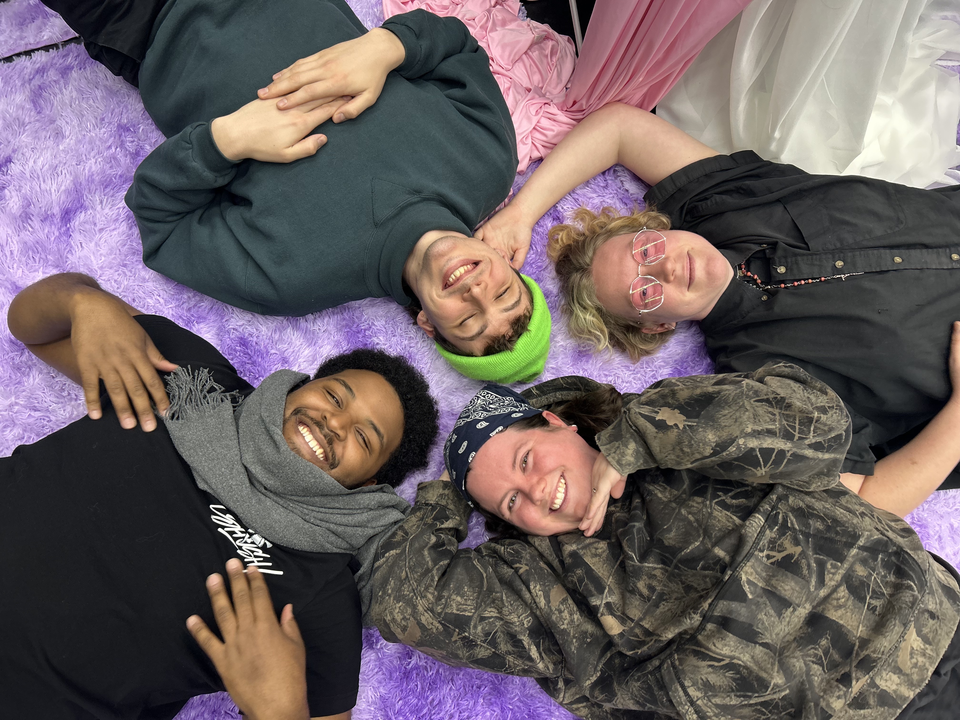 Four best friends, two white non binary folks, a Black man and a white man, lying on a bright pink fuzzy carpet in a circle looking up at the camera overhead. All smiling. All cool. All happy.